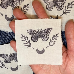 Op de afbeelding: Vier kleine, vierkante stoffen patches met een vlinderontwerp. Elke patch heeft een gedetailleerde vlinderillustratie in donkergrijs, omlijst door takjes en een halve maan. De patches liggen op een denim achtergrond.