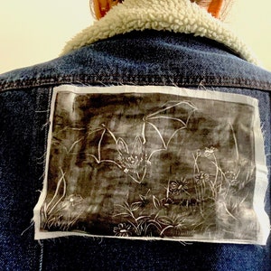 Pode incluir: Uma jaqueta de ganga azul com um remendo branco nas costas. O remendo apresenta uma ilustração em preto e branco de morcegos voando sobre um campo de flores.