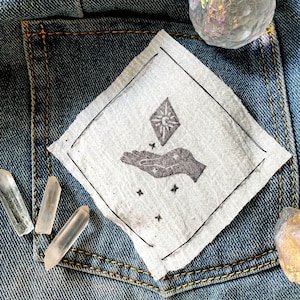 May include: A white fabric patch with a black hand holding a diamond with a sun inside. The patch is in a blue denim pocket with three clear quartz crystals and a iridescent crystal.