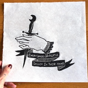 Puede incluir: Impresión en blanco y negro que representa una mano sosteniendo una daga, con el texto "EVERY LOVER'S GOT A LITTLE DAGGER IN THEIR HAND". La impresión tiene una estética vintage. Un pequeño parche de Fall Out Boy es visible.