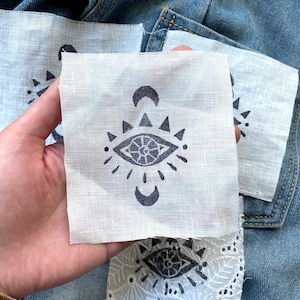 May include: Three white fabric patches featuring a black eye, crescent moon, and triangle design. The patches are displayed against a blue denim backdrop.