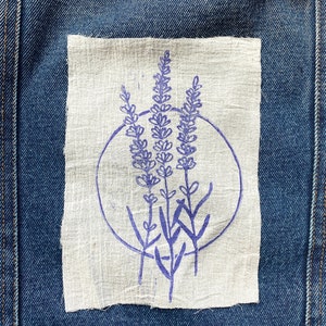 Könnte beinhalten: Ein weißer Stoff-Patch mit einem lila Lavendelblumen-Design, der auf eine blaue Jeansjacke genäht ist.