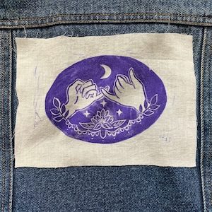 Könnte beinhalten: Ein lila und weiß gestickter Patch mit zwei ineinander verschlungenen Händen, einem Halbmond und Sternen. Der Patch befindet sich auf einem Jeansstoff.