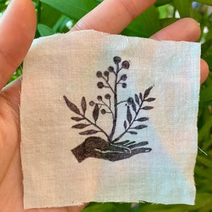 May include: A black and white illustration of a hand holding a small plant with leaves and berries. The illustration is printed on a white fabric square.