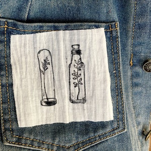 May include: Two glass bottles with black outlines, each containing a small plant, are sewn onto a white fabric patch. The patch is attached to a blue denim jacket.