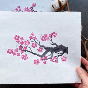 May include: A black and white print of a cherry blossom branch with pink flowers. The print is on white paper and has a simple, minimalist style.