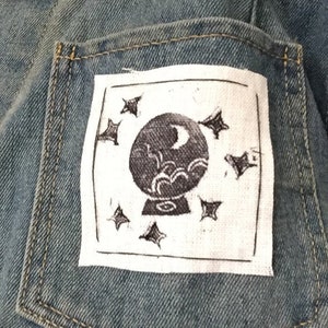 May include: A white patch with black embroidery of a crystal ball with a crescent moon and stars on a denim jacket.