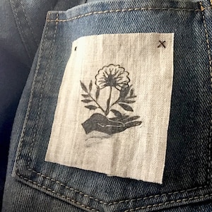 May include: A denim jacket with a patch pocket featuring a hand holding a flower with leaves. The patch is made of a light beige fabric and has a black X stitch in the top right corner.