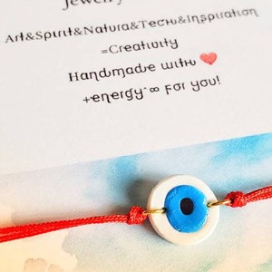 Puede incluir: Pulsera hecha a mano con un dije de ojo turco blanco y azul, ensartada en un cordón rojo. La imagen incluye texto de la marca "ARCALE" y la frase "Jewelry Design". El fondo es un lavado de acuarela.