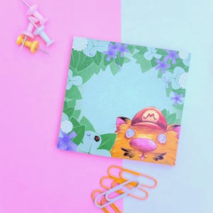 May include: A blue and green notepad with a cartoon cat wearing a red and white hat peeking out from behind a leafy border. The notepad is on a pink and blue background with yellow and pink push pins and silver paper clips.