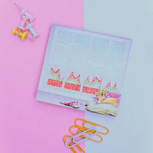 May include: A purple notepad featuring a window scene with a cat wearing a crown sleeping on a pillow. The window has a row of red flowers in pots. There are yellow and white push pins and silver paper clips on a pink and white background.