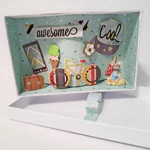 Könnte beinhalten: Eine weiße Box mit einem blauen und grünen gemusterten Innenraum. Die Box ist geöffnet und enthält eine Vielzahl von Papierschnitten, darunter ein Fahrrad, ein Koffer, eine Karte, ein Eis am Stiel, eine Maus und einen Hut mit dem Wort "cool" darauf. Die Box hat auch einen Papierschnitt des Wortes "awesome" auf der Oberseite.
