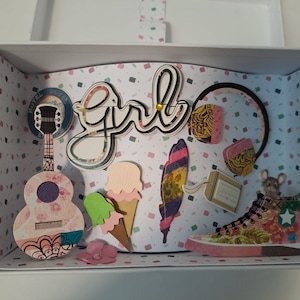 Könnte beinhalten: Eine weiße Box mit einem bunten gemusterten Hintergrund. Die Box enthält eine Vielzahl von Papierarbeiten, darunter eine Gitarre, Kopfhörer, einen Schuh, Eiswaffeln, eine Feder und das Wort "Girl" in schwarzer Schrift.