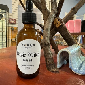 Puede incluir: Una botella de vidrio marrón con un gotero y una etiqueta blanca que dice "WYCH ELM botanicals Basic Witch BODY OIL Ingredients: sweet almond oil, ácido hialurónico, aceite de vitamina E, aceites esenciales"
