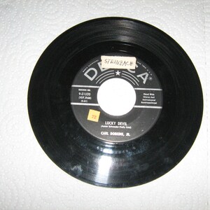 May include: A black vinyl record with a white and black label. The label reads "LUCKY DEVIL" by Carl Dorkins, Jr. The record is a 45 RPM single from Decca Records.
