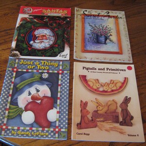 May include: Four different craft books with colourful covers. The first book is titled "Santas - Make It Easy" and features a Santa Claus with a red hat and white beard. The second book is titled "The Secret Garden - Home Decor" and features a floral arrangement in a blue watering can. The third book is titled "Just a Thing or Two" and features a snowman holding a red heart. The fourth book is titled "Pigtails and Primitives - 10 New Country Woodcraft Patterns" and features a watermelon with three bunnies on top.