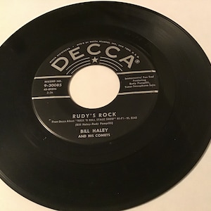 May include: A black vinyl record with a white label featuring the text "RUDY'S ROCK" by Bill Haley and His Comets. The record label also includes the Decca Records logo and the text "ROCK 'N ROLL STAGE SHOW".