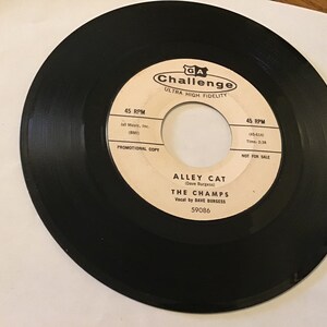 May include: A black vinyl record with a white label. The label has the text "GA Challenge Ultra High Fidelity" and "Alley Cat (Dave Burgess) The Champs Vocal by Dave Burgess 59086". The label also has the text "45 RPM" and "Promotional Copy" on the left side and "45 RPM Time: 2:28 Not for Sale" on the right side.