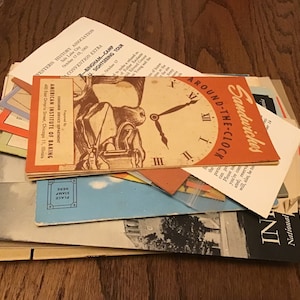May include: A collection of vintage ephemera, including brochures and postcards, is arranged on a wooden surface. The items feature text and graphics, including a clock design with the words "Sandwiches Around-the-Clock."