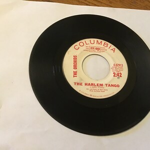 May include: A black vinyl record with a white label. The label has red text that reads "Columbia", "The Orchids", "The Harlem Tango", "A Goffin - C King", "Arr. and Cond. by Burt Kamm", "Prod. by Steve Venet", "4-42913", "JZSP 31323", "Columbia Records", "Inc.", "2:42".