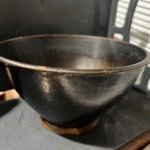 May include: A dark, rustic bowl with a rounded shape and a slightly flared rim. The bowl has a matte black finish with hints of brown and sits on a small, wooden base. This bowl is suitable for serving or display.