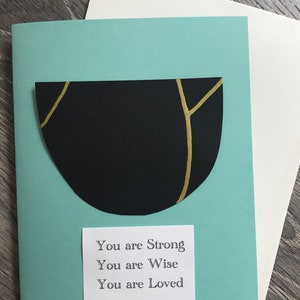 May include: A teal card with a black and gold geometric shape and a white card with the text "You are Strong, You are Wise, You are Loved" on it.