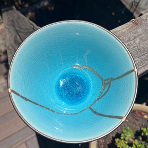May include: A round ceramic bowl with a vibrant turquoise glaze and a fine crackle pattern. The interior features a prominent gold-colored repair line running across the surface, highlighting the kintsugi technique used to mend the ceramic.