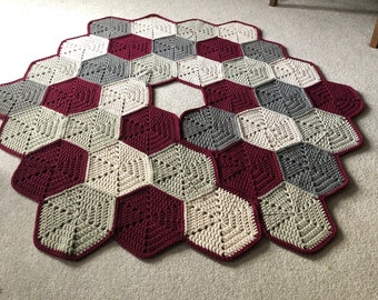 May include: A crocheted Christmas tree skirt in burgundy, beige, and gray. The skirt is made of hexagonal shapes and has a hole in the center for the tree trunk.