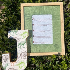 May include: A framed print with a light wood frame and a green textured background. The print has the word "JUNE" spelled out vertically with Bible verses. A large wooden letter "J" with a dinosaur pattern is next to the frame.