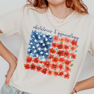 May include: Cream-colored t-shirt featuring a floral American flag design. The flag is composed of blue watercolor stars, red flowers, and the text "obstetrics & gynecology" in a script font. The shirt is a casual style.