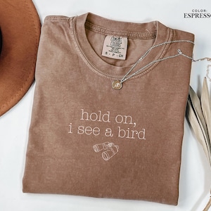 May include: Espresso-colored t-shirt with the text "hold on, i see a bird" and a graphic of binoculars. A silver necklace with a pendant is draped across the shirt. The shirt is folded, and a brown hat is partially visible.