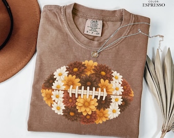 May include: Espresso-colored t-shirt with a floral football design. The football is composed of white, yellow, and brown flowers. The shirt has a crew neck and a Comfort Colors tag. A silver necklace with a small pendant is draped across the shirt.