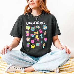 Peut inclure: Un t-shirt noir avec le texte "Back in my day..." imprim&eacute; sur le devant. Le t-shirt pr&eacute;sente un graphique color&eacute; de diverses ic&ocirc;nes de la culture pop des ann&eacute;es 90 et du d&eacute;but des ann&eacute;es 2000, notamment un Tamagotchi, une cassette audio, une Game Boy et une paire de rollers.