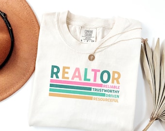 May include: Cream-colored t-shirt with the word "REALTOR" in teal and pink, above colorful horizontal stripes. Below the stripes are the words "RELIABLE, TRUSTWORTHY, DRIVEN, RESOURCEFUL" in a matching color palette. A gold necklace and a brown hat are also visible.