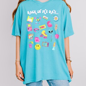 Peut inclure: Un t-shirt bleu turquoise avec le texte "Back in my day..." imprim&eacute; sur le devant. Le t-shirt pr&eacute;sente une vari&eacute;t&eacute; d'illustrations sur le th&egrave;me des ann&eacute;es 90, notamment une cassette audio, un Tamagotchi, un patin &agrave; roulettes et un Rubik's Cube.