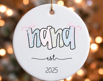 May include: White ceramic Christmas ornament with the word "nana" in a floral and striped design, with pink bows. Below the word "nana" is the word "est" and the year "2025". The ornament hangs from a gold string.