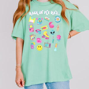 Peut inclure: Un t-shirt vert avec le texte "Back in my day..." imprim&eacute; sur le devant. Le t-shirt pr&eacute;sente une vari&eacute;t&eacute; de graphismes sur le th&egrave;me des ann&eacute;es 90, notamment une cassette audio, un Tamagotchi, un troll, un Rubik's Cube et un smiley.