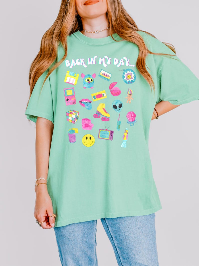 Peut inclure: Un t-shirt vert avec le texte "Back in my day..." imprim&eacute; sur le devant. Le t-shirt pr&eacute;sente une vari&eacute;t&eacute; de graphismes sur le th&egrave;me des ann&eacute;es 90, notamment une cassette audio, un Tamagotchi, un troll, un Rubik's Cube et un smiley.