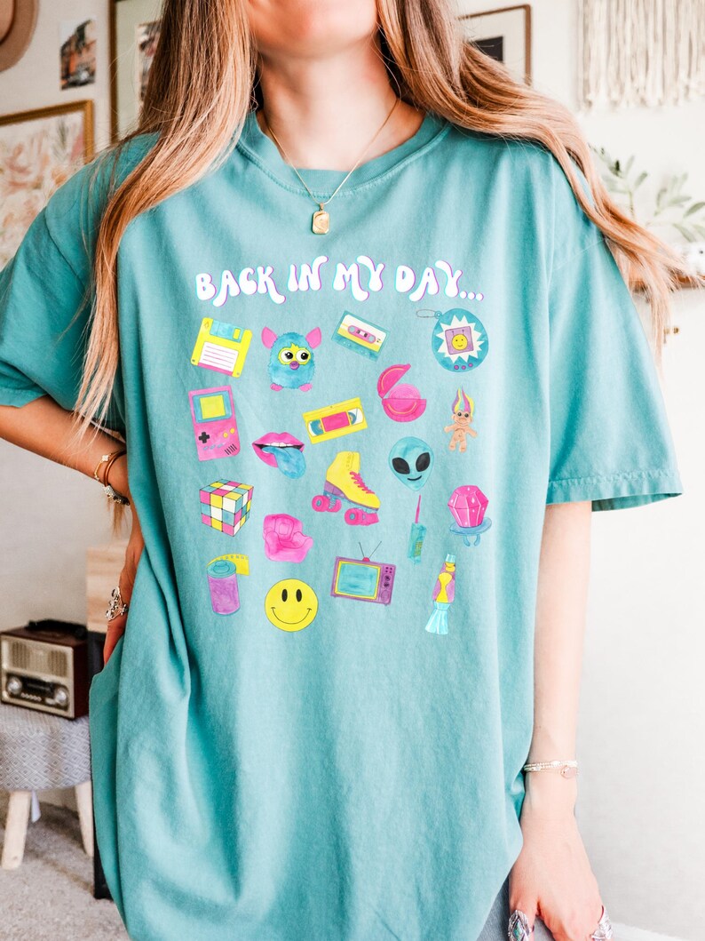 Peut inclure: Un t-shirt bleu turquoise avec un imprim&eacute; graphique de diverses ic&ocirc;nes des ann&eacute;es 90, notamment un Tamagotchi, une cassette audio, un Rubik's Cube et un smiley. Le texte "Back in my day..." est imprim&eacute; sur le t-shirt.