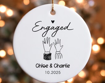 May include: A white ceramic ornament with the word "Engaged" in black script, a heart, and a drawing of two hands with a ring. Below, it reads "Chloe & Charlie 10.2025". A gold string is attached for hanging.