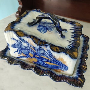 May include: A blue and gold ceramic butter dish with a lid and a handle. The dish has a floral design and a scalloped edge.