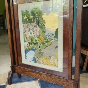 May include: A wooden fire screen with a glass panel and a framed needlepoint picture of a cottage with a river and trees. The screen has a dark brown finish and stands on four legs.