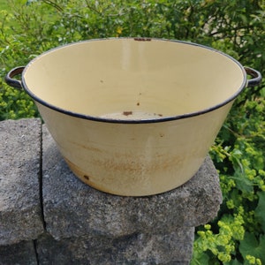 May include: A vintage enamelware basin with a cream-coloured interior and a black rim. The basin has a worn, rustic look with brown stains and rust spots.