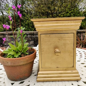 May include: A small wooden box painted gold with a single drawer and a gold knob. The box is sitting on a white table with a potted plant in the background.