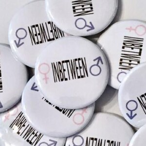 May include: A collection of white round buttons with the text "INBETWEEN" and a pink and blue gender symbol on each button.
