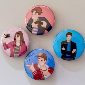 May include: Four round button pins with different illustrations of people. The first pin features a person with brown hair and a blue shirt holding a video game controller. The second pin features a person with brown hair and a red shirt taking a selfie. The third pin features a person with brown hair and a blue shirt with their arms crossed. The fourth pin features a person with brown hair and a red shirt pointing to the left and right.