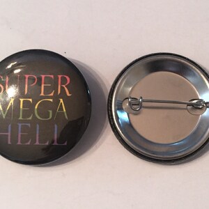 May include: A black button with a rainbow colored text that reads "SUPER MEGA HELL". The back of the button is silver with a pin.
