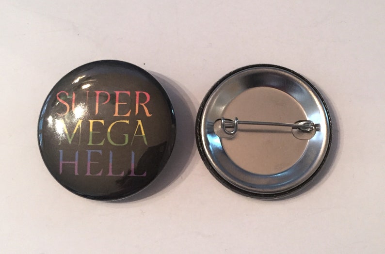 May include: A black button with a rainbow colored text that reads "SUPER MEGA HELL". The back of the button is silver with a pin.
