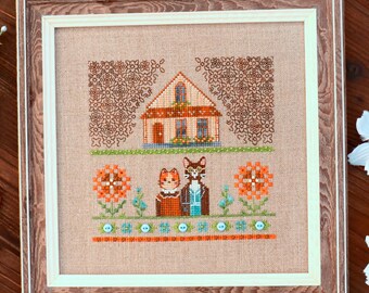 May include: Cross-stitch embroidery of a house with two cats in front of it. The house is brown with white trim and a green roof. The cats are brown and white with orange and blue accents. The background is a beige fabric with brown and orange flowers.
