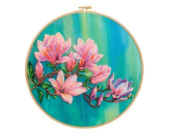 May include: A round embroidery hoop with a blue and green background featuring pink and white magnolia flowers with green leaves. The flowers are beaded and the background is a watercolour wash.